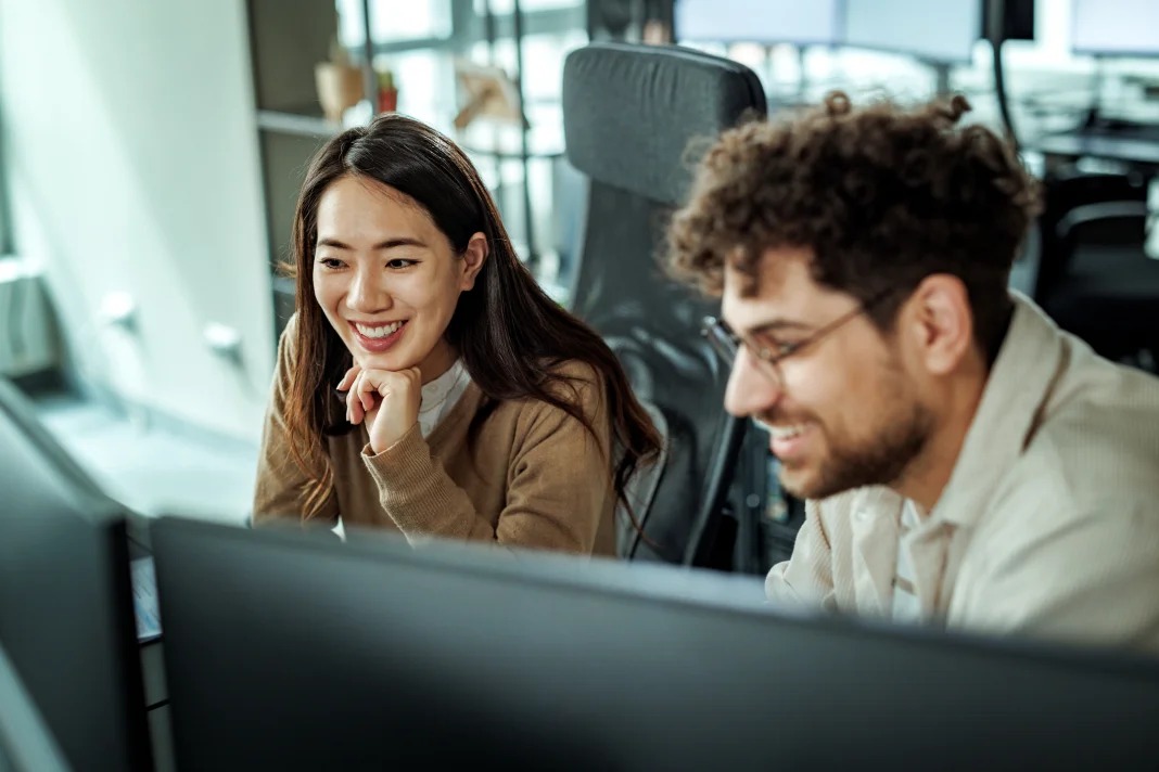 Deux personnes souriantes travaillant ensemble sur un ordinateur dans un environnement de bureau moderne.